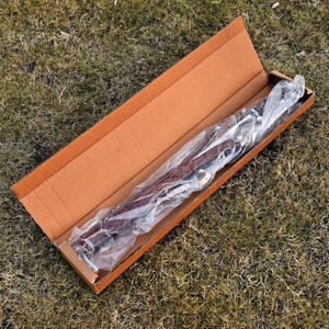 May include: A brown cardboard box, open to reveal a long, leather item wrapped in clear plastic. The leather appears to be a deep reddish-brown color, with silver-colored metal hardware visible. The box is resting on a bed of green grass.