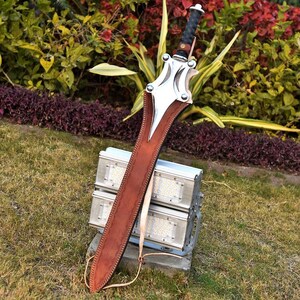 May include: A large sword with a brown leather sheath. The sword has a silver blade and guard with a black handle. The sheath is stitched with a light-colored thread. The sword is resting on a metal structure in a grassy area.