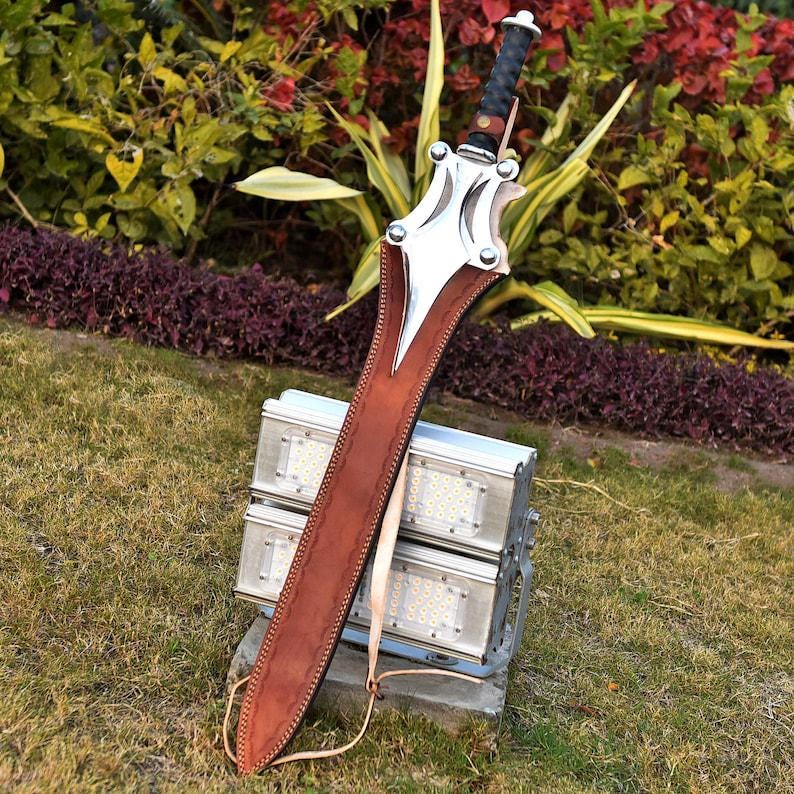 May include: A large sword with a brown leather sheath. The sword has a silver blade and guard with a black handle. The sheath is stitched with a light-colored thread. The sword is resting on a metal structure in a grassy area.
