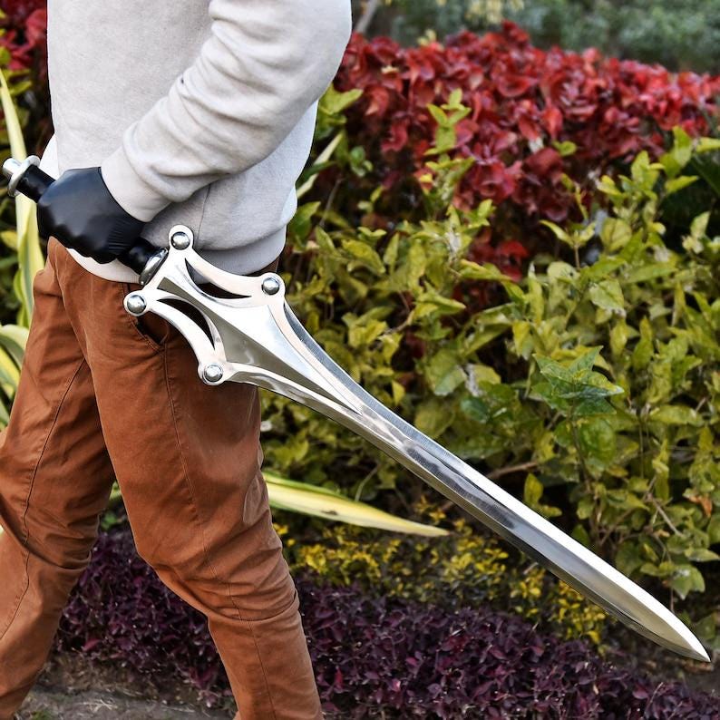 May include: A polished silver sword with a decorative hilt and a long, pointed blade. The sword is held by a person wearing a gray sweater and brown pants. The background features green and red foliage.