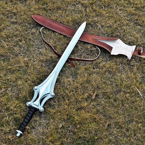 May include: A silver sword with a black handle and a decorative hilt rests on the grass next to a brown leather sheath. The sword has a long, curved blade and a detailed design on the hilt.