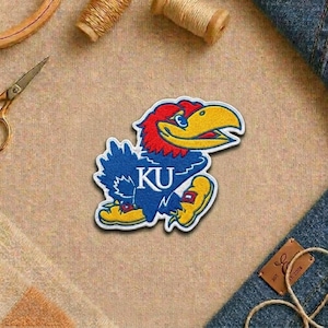May include: A colorful patch featuring a stylized blue Jayhawk mascot with red, yellow, and white accents. The patch includes the letters "KU" in white. The patch is likely for apparel or accessories.