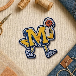 May include: Embroidered patch featuring a stylized yellow letter "M" in a basketball theme. The patch has a blue outline and depicts a basketball and a player dribbling. The patch is on a cream-colored fabric.