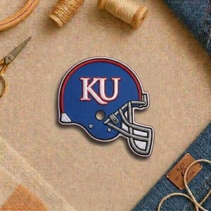 May include: Embroidered patch of a blue football helmet with the letters "KU" in white. The helmet has a red outline and silver face mask. The patch is on a tan fabric surface.
