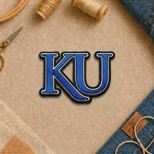 May include: Embroidered patch featuring the letters "KU" in blue with a white and black outline. The patch is on a tan fabric surface, with sewing supplies and denim accents in the background.
