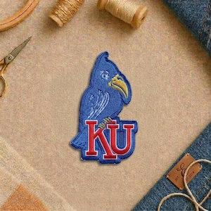 May include: A blue and red embroidered patch featuring a stylized bird and the letters "KU". The patch has a detailed design with white stitching and a yellow beak. The patch is likely for apparel or accessories.