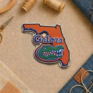 May include: Embroidered patch in the shape of Florida, with the word "Gators" in blue and white script. The patch features a green alligator head with white teeth. The background is orange with a white border.