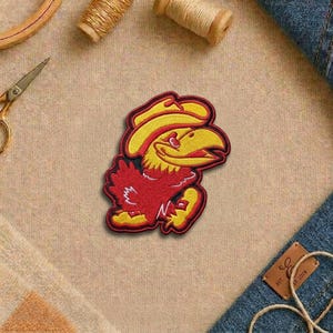 May include: Embroidered patch featuring a red and yellow cartoon bird wearing a cowboy hat. The patch has a black border and is set against a tan fabric background. The bird is smiling with its beak open.