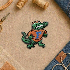 May include: Embroidered patch featuring a green alligator wearing an orange sweater with a blue "F" on the front. The alligator is wearing an orange hat and has a white-toothed grin. The patch is on a tan fabric background.