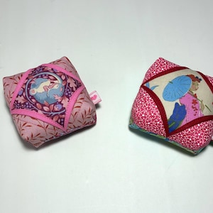May include: Two square fabric pincushions featuring folded quilt-style construction. The left cushion displays a pink floral pattern with a central circular motif of a sleeping animal. The right cushion features a pink floral print with a central panel depicting a figure holding a blue umbrella.