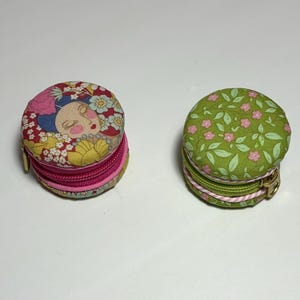 May include: Two round fabric coin purses shaped like macarons. The left purse features a colourful floral pattern with a stylised face, while the right purse has a green background with small pink flowers and leaves. Both have zips.