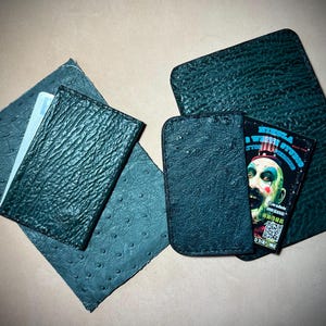 May include: Dark teal leather wallets and cardholders. One wallet is open, revealing a card with a clown graphic and text. The leather has a textured, patterned surface. A light blue card is visible in one of the cardholders.