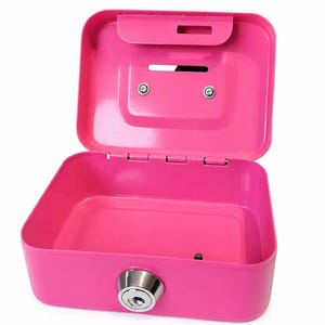 May include: A bright pink metal cash box with the lid open, revealing an empty interior. The lid features a horizontal coin slot and a recessed handle area. A circular silver-colored key lock is positioned on the front exterior panel.
