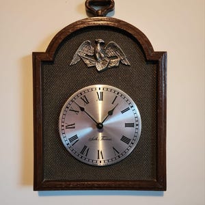 May include: A wall clock with a silver face and black hands, featuring Roman numerals. The clock is set in a dark brown wooden frame with a woven texture and a decorative metal eagle. The clock face reads "Seth Thomas".
