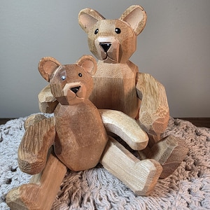 May include: Two carved wooden bear figurines in a light brown finish. The larger bear sits behind the smaller bear, with one arm resting on its shoulder. Both bears feature faceted surfaces, dark painted noses, and small black eyes.