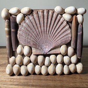 May include: A rectangular wooden frame decorated with various seashells and stones. A large, fan-shaped shell is centered, surrounded by smaller, oval-shaped stones and elongated, cylindrical shells. The color palette includes shades of brown, beige, and purple.