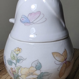 May include: White ceramic jar with a lid, decorated with pastel butterflies and floral designs. The jar has a rounded base and a lid shaped like a bird's head. The floral design includes yellow, pink, and green flowers.