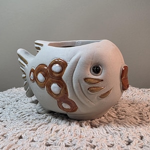 May include: A ceramic fish-shaped planter with a matte off-white finish. The design features brown and white circular patterns along the side, carved gill details, a dark eye, and a brown mouth. The top is open for planting, and the tail is visible.