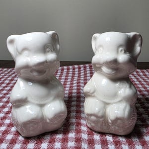 May include: Two off-white ceramic bear-shaped figurines. The bears have a smiling expression, small ears, and rounded bodies. They are sitting upright. The figurines are displayed on a red and white checkered surface.