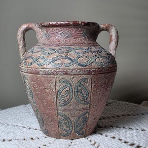 May include: A textured, antique-style vase with two handles. The vase features a reddish-brown base color with blue and cream-colored decorative patterns. The rim is a darker red. The vase is sitting on a white, crocheted surface.