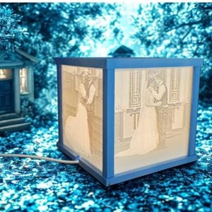May include: A blue-framed, illuminated box featuring a wedding scene. The box displays a couple in a wedding dress and suit. The box is connected to a power cord and sits in a blue-toned outdoor setting with a miniature house in the background.