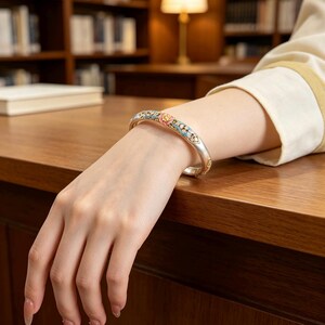 May include: A silver-toned metal bangle bracelet featuring intricate enamel detailing in shades of pink, light blue, and gold. The bracelet has a smooth, polished finish and a curved, hinged design that rests securely around the wrist.