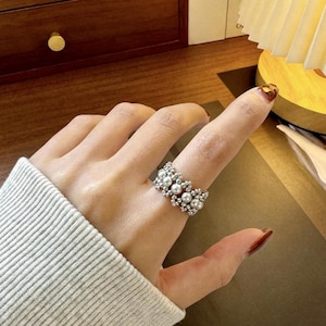 May include: A wide ring featuring a woven pattern of small silver-toned beads and white spherical faux pearls. The band has a textured, multi-layered appearance created by the alternating arrangement of the metallic beads and the smooth white pearls.