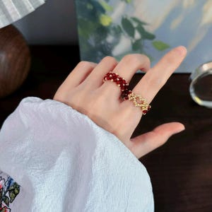 May include: A ring featuring a band of small, deep red faceted beads interspersed with tiny gold-toned metallic beads. The design incorporates a central heart-shaped motif crafted from the same gold-toned beads, creating a textured, beaded appearance.