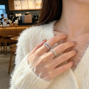 Op de afbeelding: Twee zilverkleurige ringen gedragen aan de vingers. De bovenste ring heeft een centrale rij lichtpaarse rechthoekige stenen geflankeerd door twee rijen kleine heldere kristallen. De onderste ring is een eenvoudige band met één rij heldere kristallen.