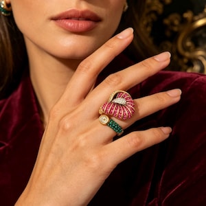 Puede incluir: Un anillo de oro con una sección superior grande en forma de abanico incrustada con filas de piedras rosas vibrantes y un pequeño grupo de piedras blancas. La banda está dividida, con una sección inferior que presenta piedras verde oscuro.