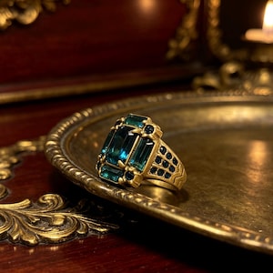 May include: A gold-toned ring featuring a large, rectangular, emerald-green central gemstone surrounded by smaller, matching green stones. The band is decorated with a series of tiny, dark blue circular stones along the sides and a textured pattern.