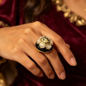 May include: A gold-toned statement ring featuring a central oval stone with a black and white spotted pattern. The centre stone is surrounded by a floral-shaped halo of six smooth, milky white cabochon stones and a black enamel base band.