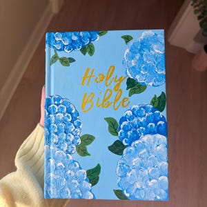 May include: A light blue book cover with hand-painted blue hydrangea flowers and green leaves. The words "Holy Bible" are written in gold script across the cover.