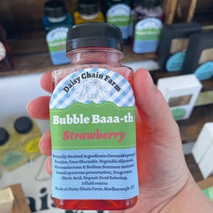 May include: A clear plastic bottle filled with red liquid, featuring a label with a blue gingham border, a goat illustration, and the text Daisy Chain Farm, Bubble Baaa-th, Strawberry, and a list of ingredients including 8 fluid ounces.