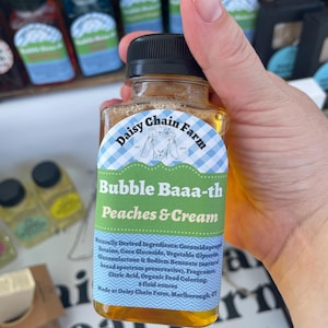 May include: A clear plastic bottle containing amber-colored liquid with a black cap. The label features a blue and white gingham border, a goat illustration, and the text Daisy Chain Farm, Bubble Baaa-th, Peaches & Cream, and 8 fluid ounces.