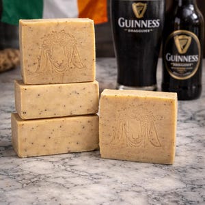 May include: Four rectangular, light brown soaps with a decorative design are stacked and arranged on a marble surface. A pint glass and a bottle of Guinness stout are in the background, along with an Irish flag.