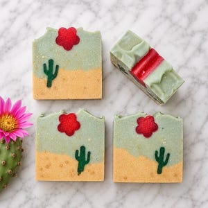 May include: Three rectangular bars of soap and one end piece, each featuring a desert landscape design with a light green sky, a tan sandy base, a dark green saguaro cactus, and a red flower. A bright pink cactus flower is positioned nearby.