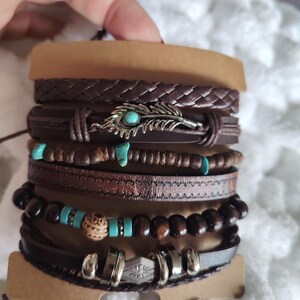 Può includere: Un gruppo di braccialetti in pelle marrone con vari design. Alcuni braccialetti presentano perline turchesi, perline di legno e un ciondolo a forma di piuma d'argento. I braccialetti sono tenuti insieme da un cartoncino marrone.