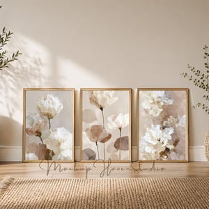 May include: Three framed floral art prints with gold frames. The artwork features abstract white and beige flowers with brown and beige accents. The prints are displayed on a wooden floor, leaning against a beige wall.