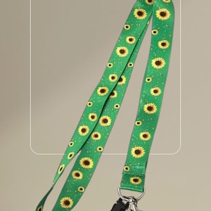 May include: Green lanyard with a repeating pattern of yellow sunflowers. The lanyard has a black plastic clasp and a silver metal clip for attaching to keys or an ID badge. The background is a solid, neutral color.