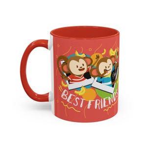 Puede incluir: Taza de cerámica blanca con interior y asa rojos. La taza presenta una ilustración de dibujos animados de dos monos en marcos de fotos, con las palabras "BEST FRIENDS" en blanco. Plátanos y círculos naranjas decoran el fondo rojo.