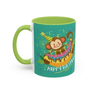 Puede incluir: Taza de cerámica con asa e interior verdes. El exterior presenta un mono de dibujos animados con gorro de fiesta, montado en un plátano con glaseado y las palabras "HAPPY BIRTHAY". La taza es blanca con fondo turquesa.