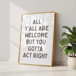 May include: A framed art print with the text "ALL Y'ALL ARE WELCOME BUT YOU GOTTA ACT RIGHT" in gray letters. The frame is gold, and the print is leaning against a white wall. A potted plant is visible in the background.