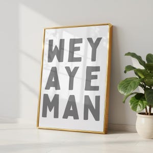 May include: A framed art print with the words "WEY AYE MAN" in gray block letters on a white background. The frame is gold, and the print is leaning against a white wall. A potted plant is in the background.