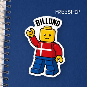 May include: A die-cut sticker featuring a yellow plastic toy figure with a smiling face, wearing a red torso with a white cross design and blue legs. The word BILLUND is printed in black above the figure's head on a white background.