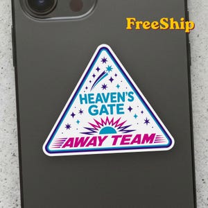 May include: A triangular sticker with a white background, blue border, and the text "HEAVEN'S GATE" in blue and "AWAY TEAM" in pink. The design features purple and blue stars, a shooting star, and a stylized sunburst graphic in pink and blue.