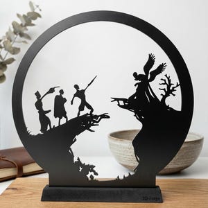 Könnte beinhalten: Schwarzes Metallkunstwerk, das Silhouetten in einem kreisförmigen Rahmen zeigt. Das Kunstwerk zeigt Figuren auf einer Klippe, die einer geflügelten Figur gegenüberstehen. Die Basis lautet "Handcrafted, Finite Art Piece."