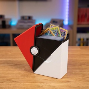 May include: A red, black and white plastic card storage box designed like a spherical toy. The box features a hinged lid that is partially open, revealing a collection of rectangular trading cards stored inside with clear protective sleeves.