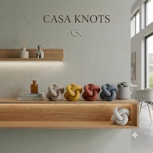 Può includere: Una collezione di sculture a nodo decorative in beige, giallo, terracotta, blu e bianco. Il testo CASA KNOTS e un logo appaiono sopra mensole in legno con un diffusore, una statuetta, una bottiglia di vetro marrone, libri e una piccola pianta.