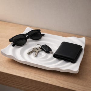 Può includere: Un vassoio bianco con bordi ondulati contiene occhiali da sole neri, chiavi, un portachiavi per auto e un portafoglio nero. Il vassoio ha un design circolare strutturato al centro. Gli oggetti sono disposti su una superficie di legno chiaro.